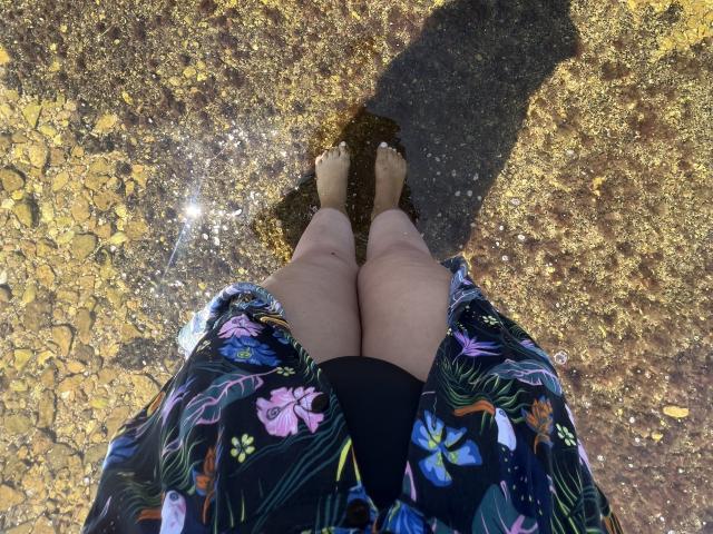 Barbarica's feet in floral kimono, bare legs, standing on wet, rocky ground with small water reflections, shadow on bottom right. Bare feet, slightly dirty, in floral pattern.