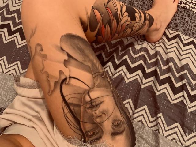 AliviyaTail's tattooed arm with a detailed black-and-white portrait of a woman's face on the upper arm, extending to a colorful flower tattoo on the forearm. She's on a zigzag-patterned bed.
