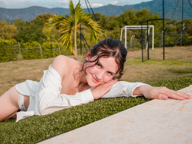 ChloeDulce sdraiata sull'erba, indossa una camicia bianca, con il seno scoperto, sorride alla fotocamera, un braccio esteso, prato verde, porta da calcio e montagne sullo sfondo..