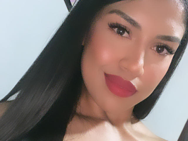 StephanieDavies with long black hair, red lipstick, and eyeliner, smiling at the camera, showing her bare shoulders and a subtle hint of her tits.