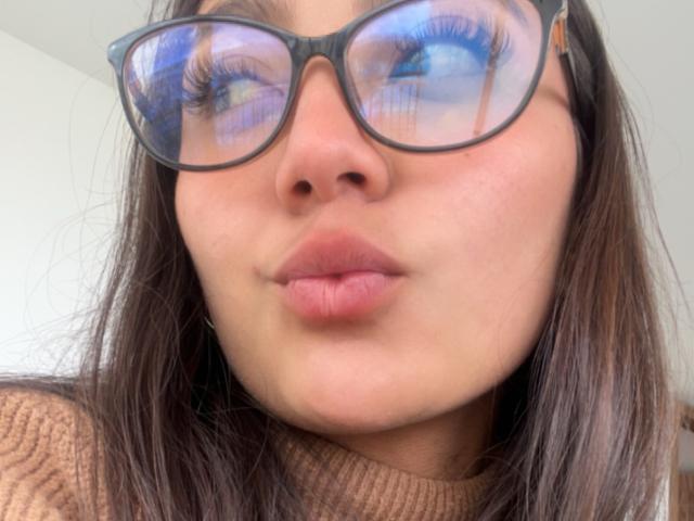 DianaFernandezz heeft volle, roze lippen, een medium bruine huid en draagt dikke, zwarte brillen met reflecterende, blauwe ogen. Ze draagt een bruine, geribbelde trui en heeft recht, bruin haar..