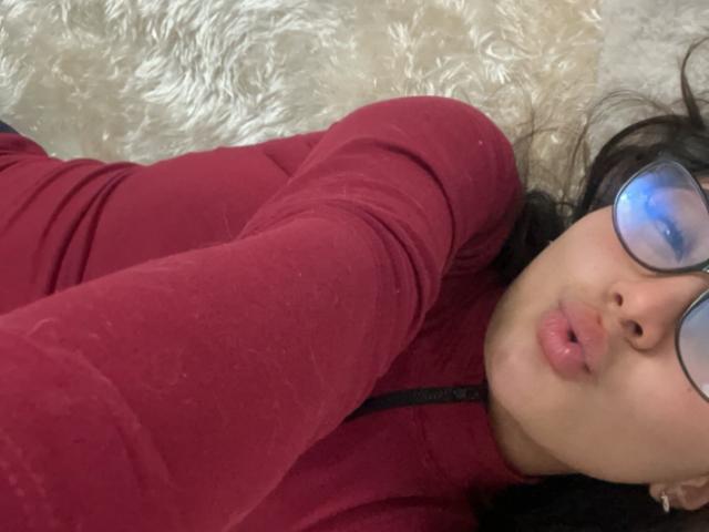 DianaFernandezz, con los labios fruncidos, vistiendo una blusa roja de manga larga y gafas, acostada sobre una superficie blanca y esponjosa, con su cabello negro ligeramente despeinado..