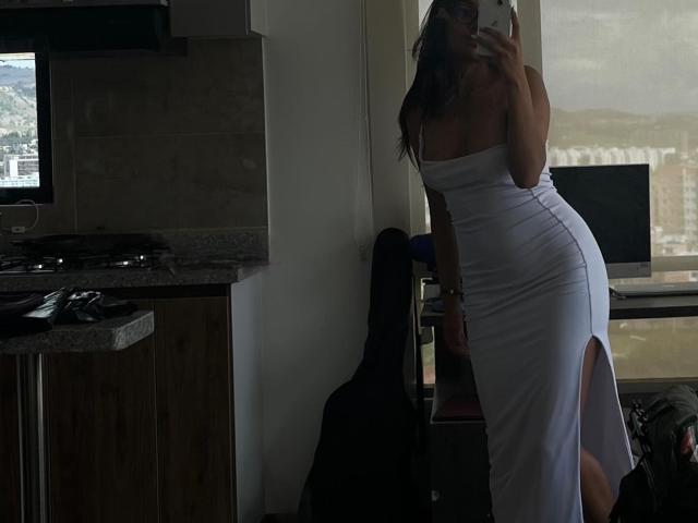DianaFernandezz, avec ses longs cheveux noirs, portant une robe blanche moulante sans bretelles, qui révèle ses fesses et une fente sur le côté, se prend en photo devant un miroir dans une cuisine peu éclairée..