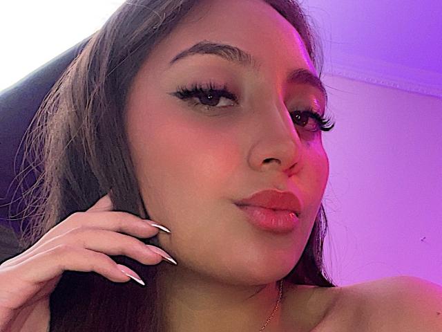 DianaFernandezz has a cute face with full lips, long dark hair, and thick eyelashes. Her hand with black and white nails touches her head. Pink and purple lighting.