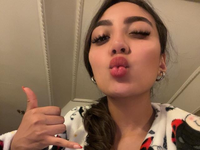 DianaFernandezz with pouty pink lips, winking, and giving a thumbs-up. She's wearing a white, red, and black patterned robe, with hoop earrings and long, curly hair.