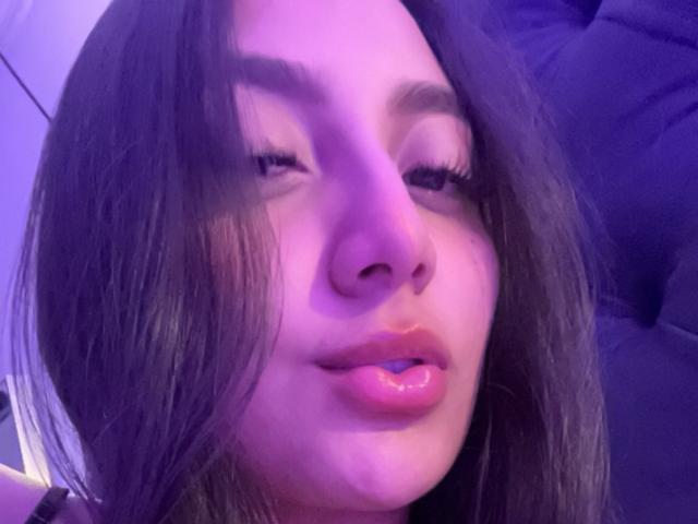 Het gezicht van DianaFernandezz, met lang bruin haar, huid met paarse tinten, licht open glanzende lippen en zichtbare blauwe lippenstift, ogen half gesloten, in een ontspannen, sensuele uitdrukking..