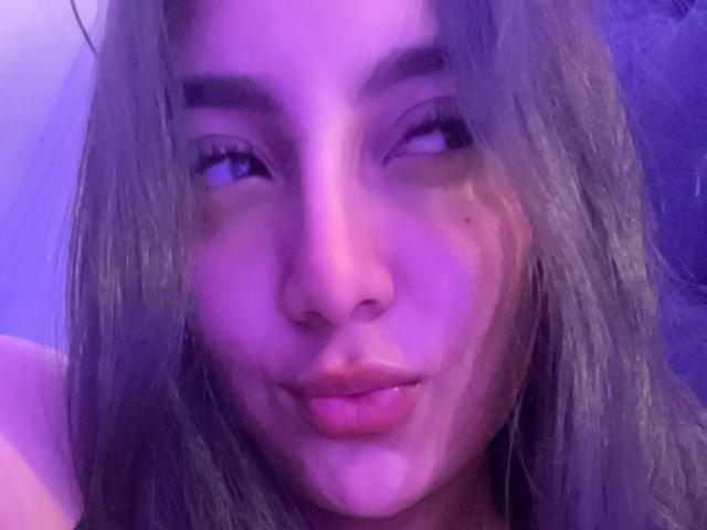 DianaFernandezz's face with purple lighting, looking sideways with pouty lips, long brown hair, and a small amount of white fluid on her chin.
