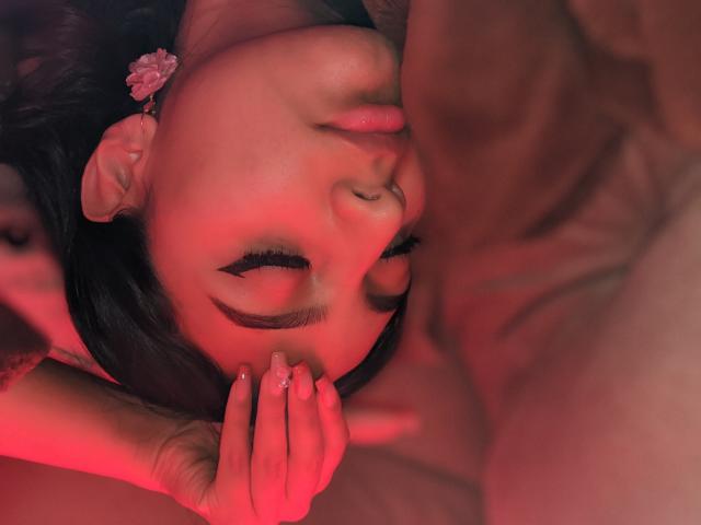 DianaFernandezz with closed eyes, black hair, and pink flower earring, sucks a large, erect penis. Her hand with a ring rests on her chin. Red lighting.