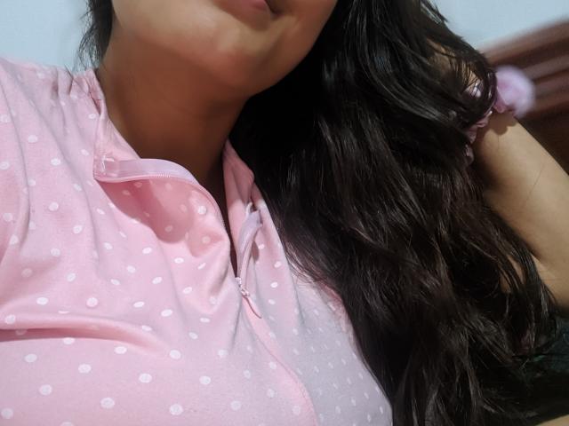 DianaFernandezz's face and upper body, wearing a pink polka dot shirt, with long dark wavy hair, partially visible pout, and right hand behind her head.