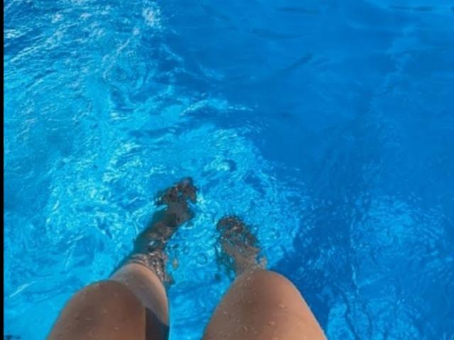 DianaFernandezz's legs in a bright blue pool, water up to her thighs, showing her brown skin and black swim fins, from a bottom-up perspective.