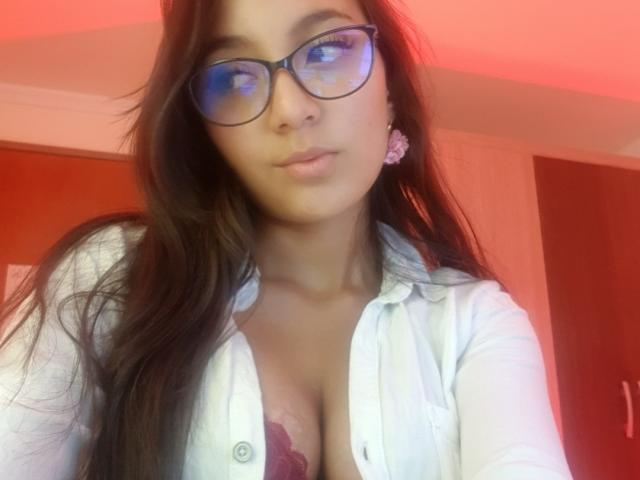 DianaFernandezz with glasses, long brown hair, white shirt open showing maroon lace bra, pink earrings, red light, soft expression, and a slight pout. Boobs visible, looking cute.