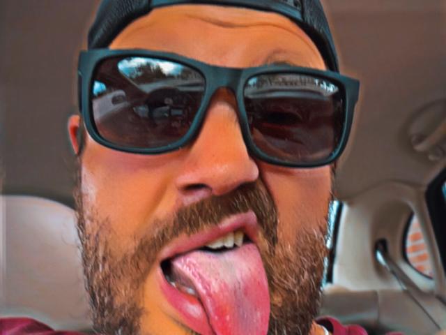 LordAdonison, con la barba, gli occhiali da sole neri e un cappello da baseball indossato al contrario, mostra una lingua umida e rosa. Il suo viso è al centro dell'immagine, con un'espressione buffa..