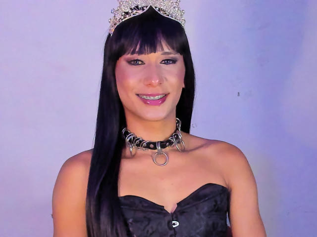MoonRedest, avec ses longs cheveux noirs, portant une tiare en argent, un corset noir sans bretelles et un collier à pointes, souriant avec des dents métalliques, a un visage mignon et de petites seins..