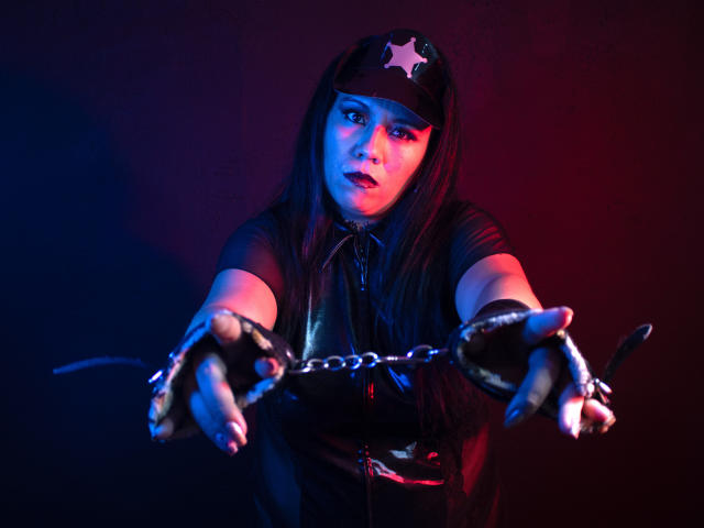 LexyDom with long black hair, wearing a police hat and black leather outfit, extends chained hands towards the camera, illuminated by blue and red lights.