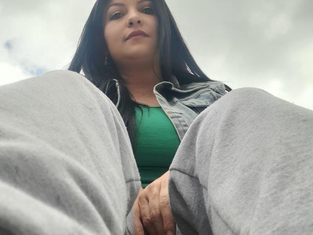 NatalyHami, con i suoi lunghi capelli neri, la maglietta verde e la giacca di jeans, è seduta con le gambe divaricate, mostrando dei pantaloni grigi, e con le mani appoggiate sulle ginocchia, vista da sotto, mentre guarda verso il basso..