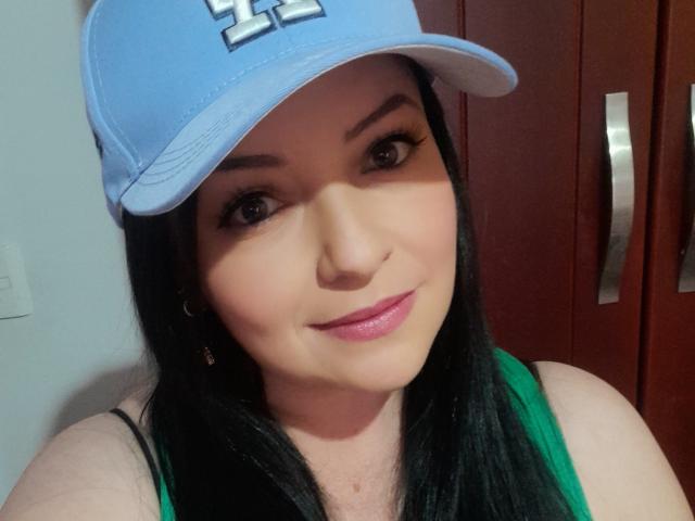 NatalyHami with black hair, wearing a blue cap, green top, and pink lipstick, smiling at the camera, showing her boobs slightly, brown eyes, and black eyeliner.