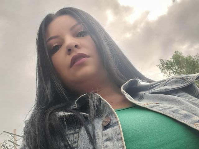 NatalyHami, avec ses longs cheveux noirs, son haut vert et sa veste en jean, regarde vers le bas avec un léger sourire. Le ciel est nuageux, et un arbre est visible sur la droite. Ses seins sont légèrement visibles..