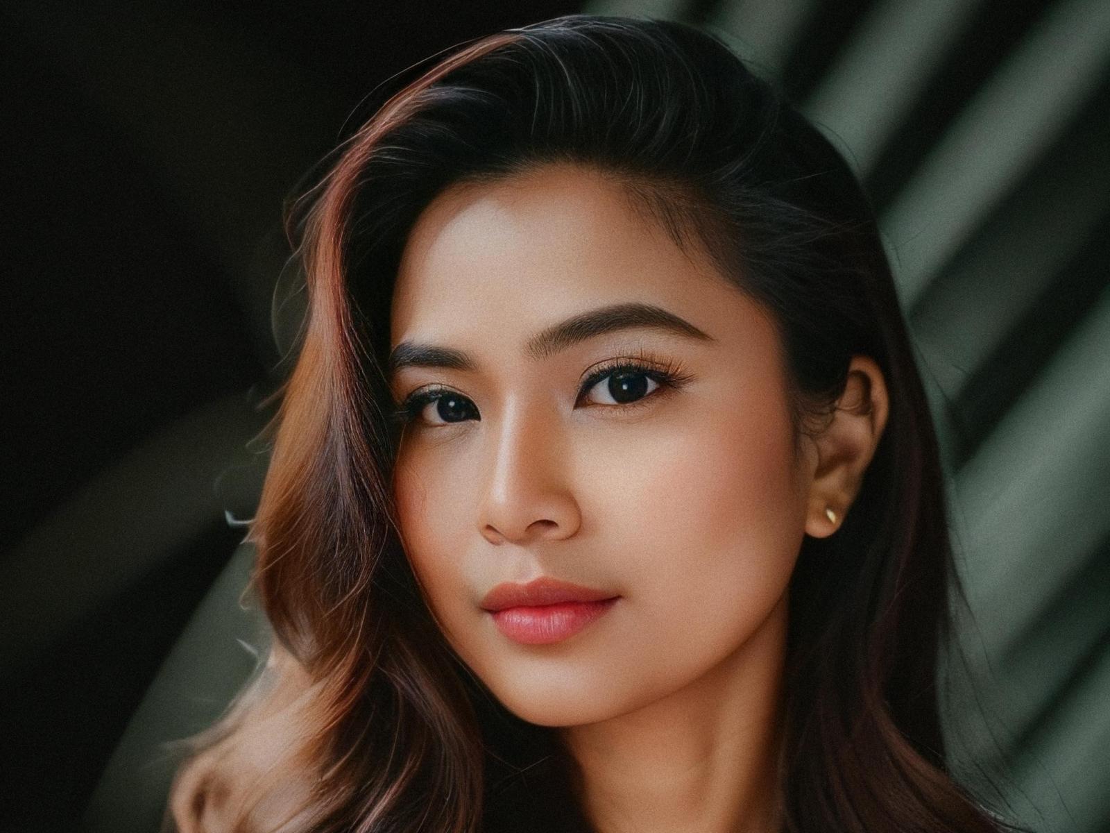 PinayPilya has smooth, tan skin, dark brown wavy hair, and a cute face with blue eyes and pink lips. She wears small gold earrings and looks directly at the camera.