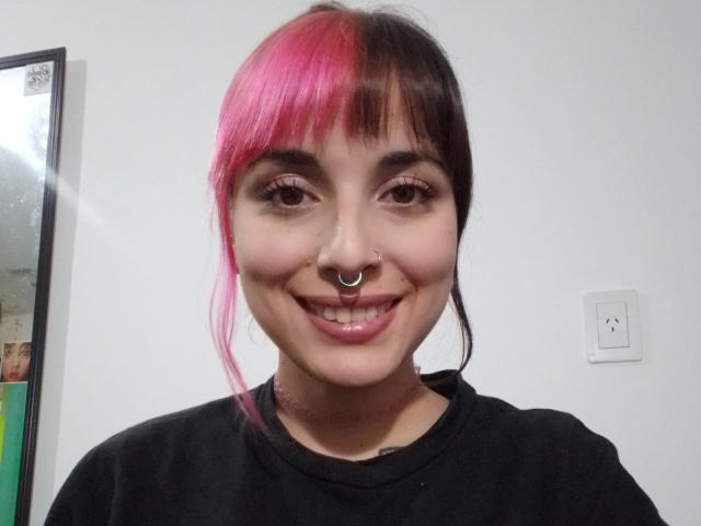 A SoyLuz tem cabelo rosa e preto, um piercing no nariz e está sorrindo com lábios rosados e brilhantes. Ela está vestindo uma camiseta preta e tem uma tatuagem no pescoço..