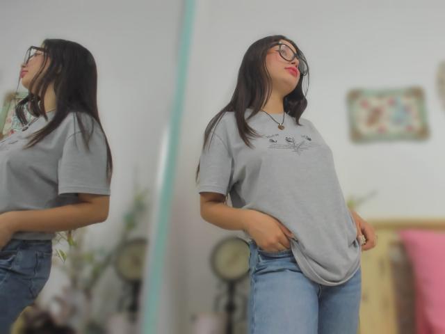 Genesiss with long black hair, glasses, and red lipstick, wearing a loose gray tee and blue jeans, standing with hands in pockets, reflected in a mirror.