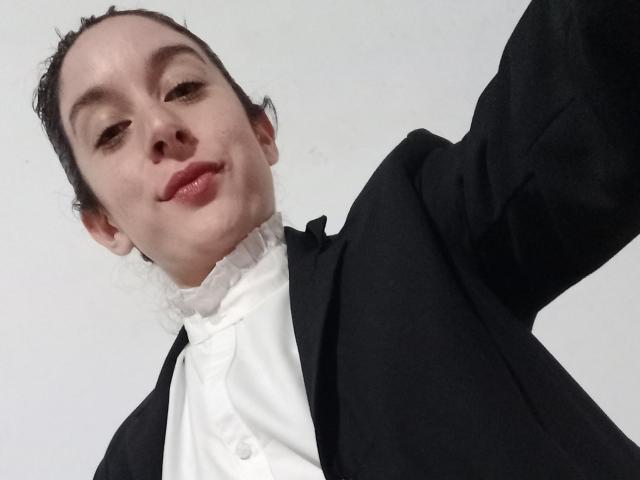 Semda a un léger sourire, les cheveux noirs attachés en chignon, et elle porte une veste noire par-dessus une chemise blanche à volants. Ses lèvres rosées sont légèrement brillantes. Elle prend un autoportrait..