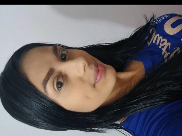 AnaisHilton has black hair, brown eyes, and pink lips, wearing a blue shirt. Her face is slightly angled, with a gentle smile and subtle makeup.