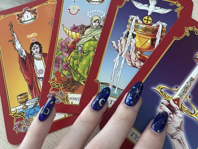 La main de Valissia, avec ses ongles bleu foncé et pailletés, tient un jeu de quatre cartes de Tarot présentant des thèmes angéliques et religieux. Les cartes sont ornées, bordées de rouge et très détaillées..