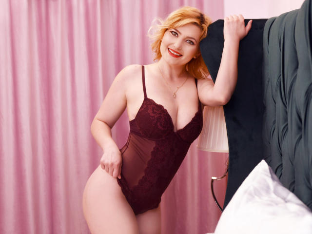 LiaPonti with blonde hair, wearing a sheer burgundy lace bodysuit, has her tits pushed up, smiling, standing against a black headboard, hand on it, red lipstick.