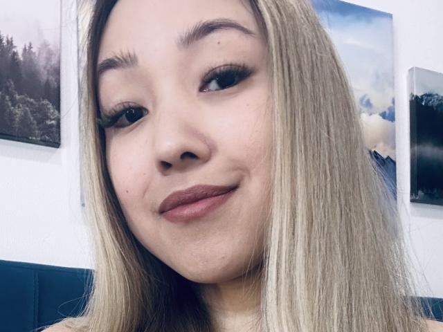 RenyLime tem a pele clara, cabelos longos e loiros, e está sorrindo, usando batom escuro e cílios postiços. Ela tem uma pequena marca de beleza abaixo do olho direito..