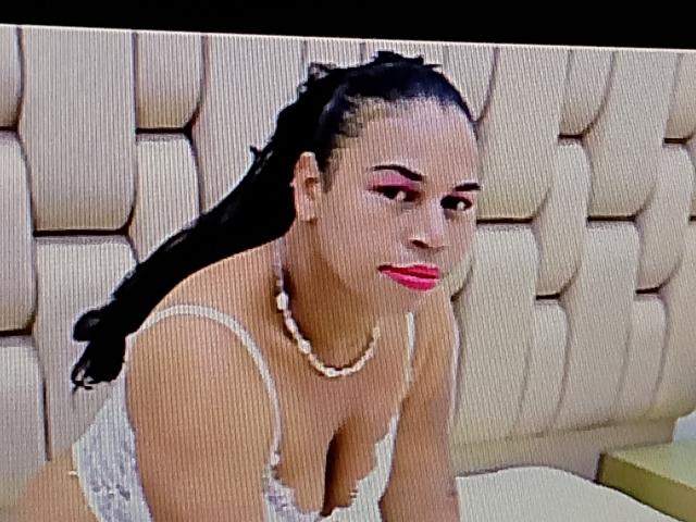 EbonnyOolsson with big boobs, black braided hair, pink eye makeup, red lipstick, white lace top, pearl necklace, leaning forward in a padded room.