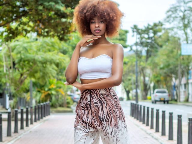AriannyOrtega with big brown afro, wearing white strapless top and zebra-patterned pants, standing on sidewalk, hand near chin, looking at camera, ass covered, background blurry.
