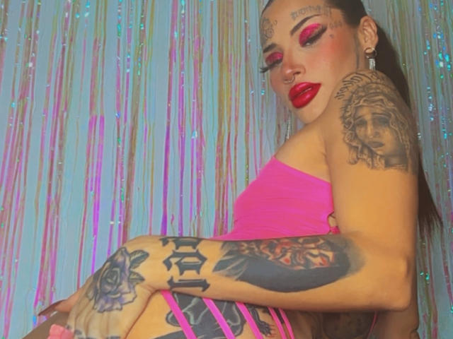 LovelyWillow, con un maquillaje rosa brillante, lápiz labial rojo y tatuajes, vestida con un top rosa sin tirantes, mostrando tatuajes en su brazo y costado, sentada con la espalda hacia la cámara..