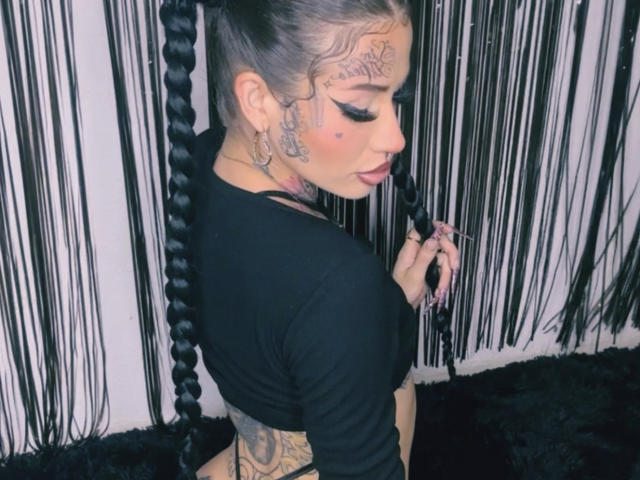 LovelyWillow con cabello trenzado, camiseta negra de manga larga, tatuajes en la cara y el brazo, sosteniendo el trenzado, con aros, mirando hacia abajo, lápiz labial rosa, fondo negro, suelo negro y esponjoso..