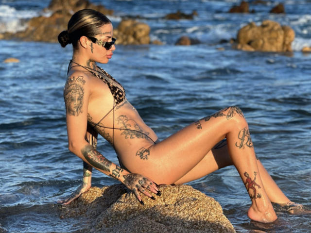 LovelyWillow, con tatuajes, cabello castaño recogido en un moño, usando gafas de sol y un bikini de estampado de leopardo, está sentada sobre una roca en el mar azul poco profundo, con las piernas cruzadas, mostrando sus pechos y glúteos..