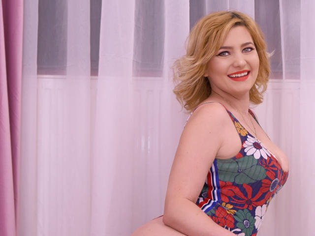 LiaPonti with blonde hair, smiling, wearing a colorful floral one-piece, showing her big boobs, standing against pink and white curtains, looking over her shoulder.