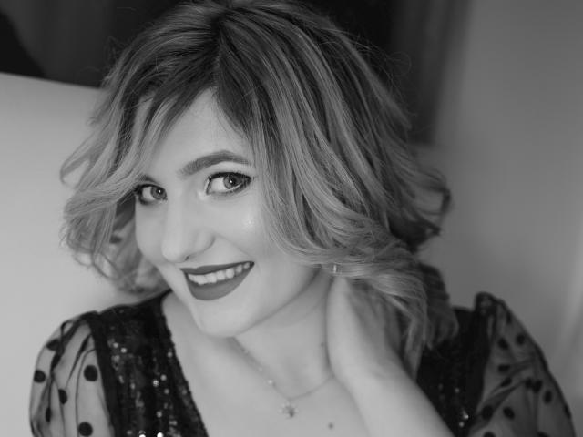 LiaPonti with short, wavy blonde hair, smiling, wearing dark lipstick, and a sparkly black top, holding her neck, in a black-and-white photo.