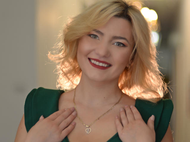LiaPonti with blonde hair, smiling, wearing a green dress, gold necklace, and red lipstick, holding her dress with her hands. Her tits are visible but not the focus.