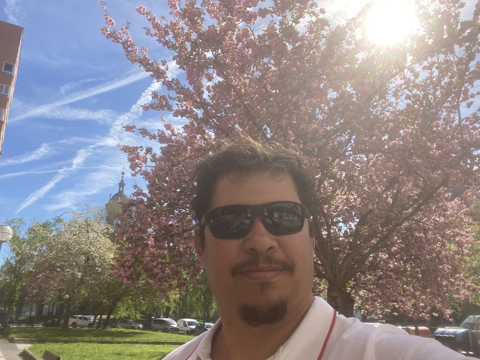 MaxSpanish has a brown mustache, small goatee, and is wearing black sunglasses. He's sporting a white shirt with a red line and is standing in front of a pink tree.