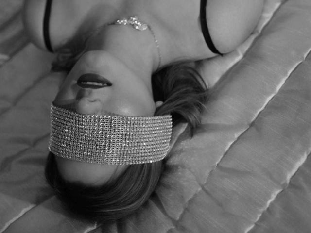 LiaPonti has her face covered by a sparkly blindfold, lying on a bed with her head tilted back, showing dark lipstick and a necklace, wearing a black bra.