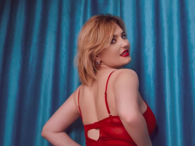 LiaPonti with blonde hair, red lingerie, and red lipstick, looking over her shoulder, showing her back and small cutout, standing in front of blue curtains.