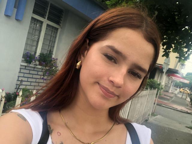 Melody Mansson tiene el pelo castaño, una nariz pequeña y una sonrisa discreta. Lleva una blusa blanca, aretes dorados y un collar, y tiene tatuajes en el pecho..