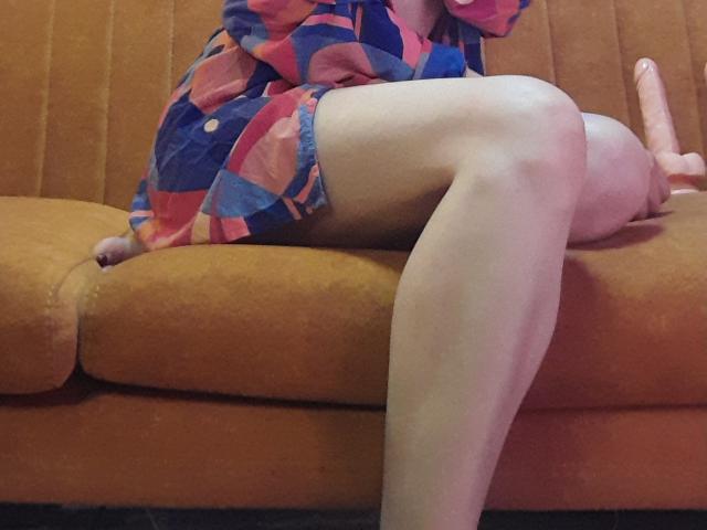 AdelaShery saß mit verschränkten Beinen auf einem braunen Sofa, trug ein farbenfrohes Kleid und hatte einen pinken Dildo zwischen ihren Oberschenkeln. Ihre helle Haut bildet einen Kontrast zum lebhaften Stoff..