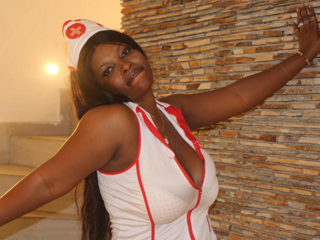 Annykaa, à la peau foncée et aux longs cheveux noirs, portant une tenue de nurse blanche avec des détails rouges, dévoilant sa poitrine généreuse grâce à un décolleté plongeant, appuyée contre un mur de pierre..