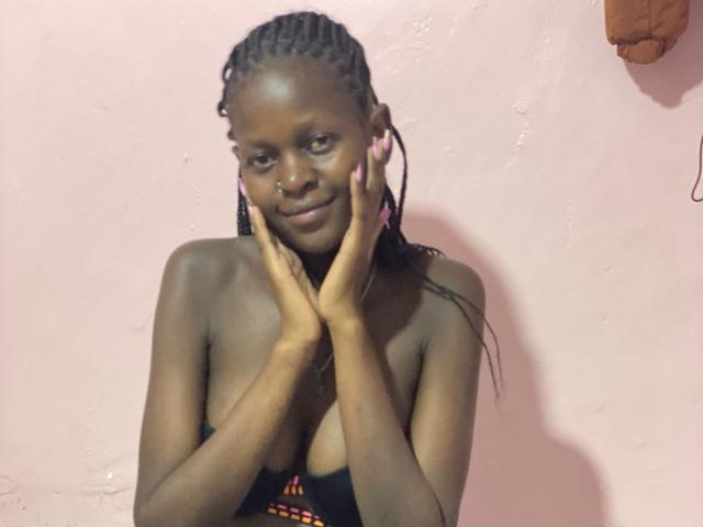 HotMitchell with dark skin, braided hair, small nose ring, smiling, hands on cheeks, topless, showing her tits, pink wall in background, brown hanging object to the right.
