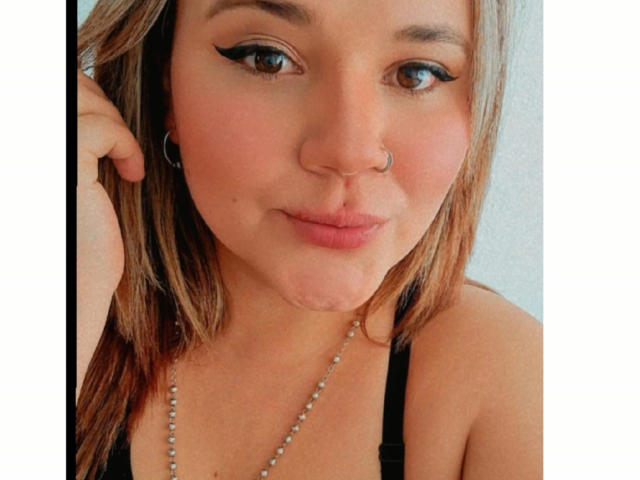 NatalieRey met zwarte eyeliner, een neusring en lichtbruin haar, lichtjes fronsend, draagt een zwarte top en een kralenketting, en kijkt met een subtiele glimlach naar de camera..