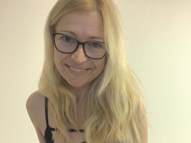 ValiaV has blonde hair, glasses, and a cute smile, wearing a black bra, with visible breasts and a slender build, in a simple, light-colored background.