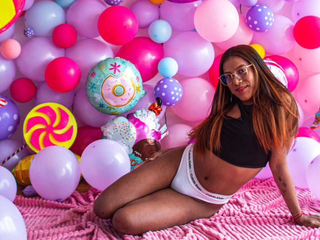 KimmySolange with long brown hair, glasses, black top, white panties, sitting among colorful balloons and candy-themed decorations, smiling, with her ass and pussy slightly visible, on pink fabric.