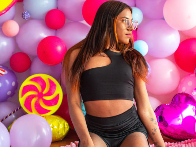 KimmySolange with long brown hair, glasses, black crop top, and black shorts, sitting among colorful balloons. Her ass is visible, and she has tattoos on her right arm.