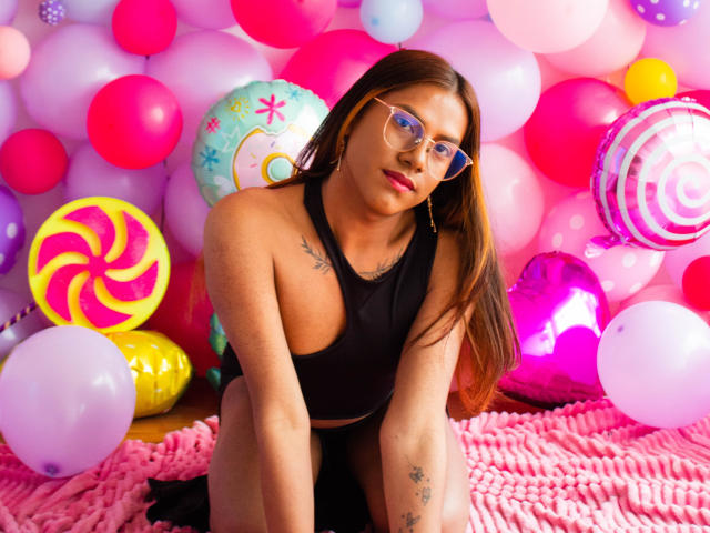 KimmySolange with long brown hair, glasses, black tank top, and tattoos, kneeling on pink blanket, surrounded by colorful balloons, showing her cute face and small tits.