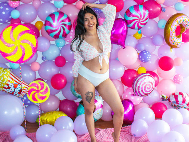 Isabella: Principessa con seno di media grandezza, biancheria intima bianca e una maglietta rosa trasparente annodata, in piedi tra palloncini colorati, sorridente, con una mano tra i capelli, che mostra un tatuaggio sulla coscia destra..